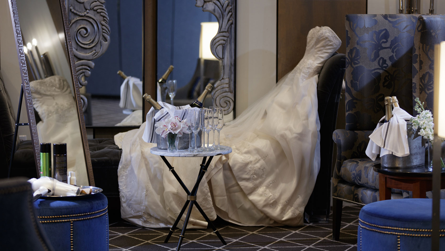 Guestroom set up to get ready for a wedding. A table with champagne and glasses sits in the middle, a wedding dress out of focus behind